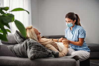 senior woman lying down on sofa while nurse with protective face mask sitting next to her and holding her hands