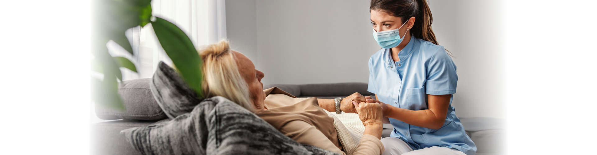 senior woman lying down on sofa while nurse with protective face mask sitting next to her and holding her hands